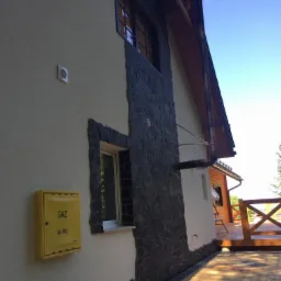 Elewacja domu z fragmentami wykończonymi kamieniem dekoracyjnym, żółta skrzynka gazowa na ścianie, widok na drewniany taras z balustradą.