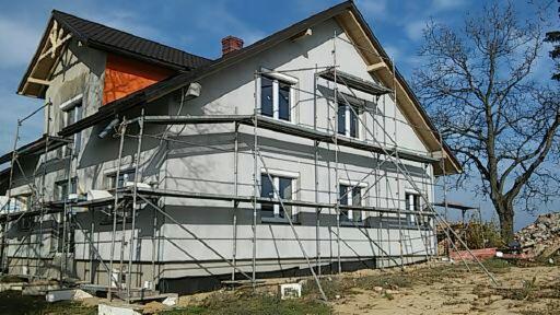 Dom jednorodzinny w trakcie wykonywania elewacji, widoczne rusztowania i fragment ocieplonego poddasza z pomarańczową wełną mineralną.