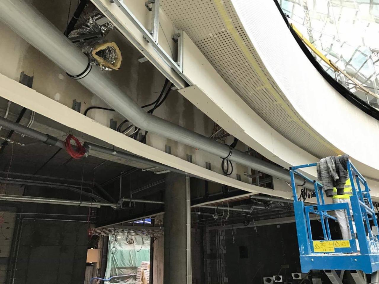 Wnętrze w trakcie wykańczania: widoczne rury wentylacyjne, kable i konstrukcja sufitu podwieszanego, pracownik na podnośniku koszowym.