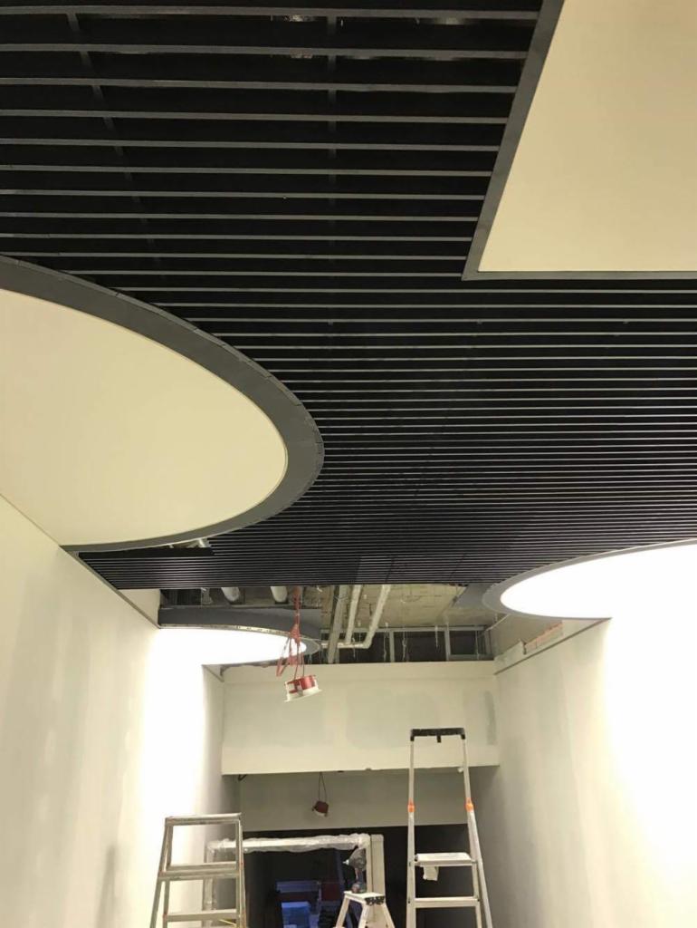 Sufit biurowy z czarnymi listwami rastrowymi i okrągłymi oprawami oświetleniowymi, widoczne drabiny i elementy instalacji w tle, wskazujące na trwające prace wykończeniowe.