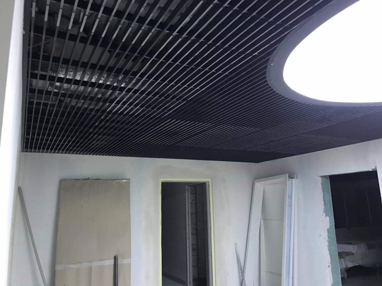 Montaż czarnego, rastrowego sufitu podwieszanego z okrągłym oświetleniem w pomieszczeniu w trakcie prac wykończeniowych, widoczne niedomalowane ściany i futryny.