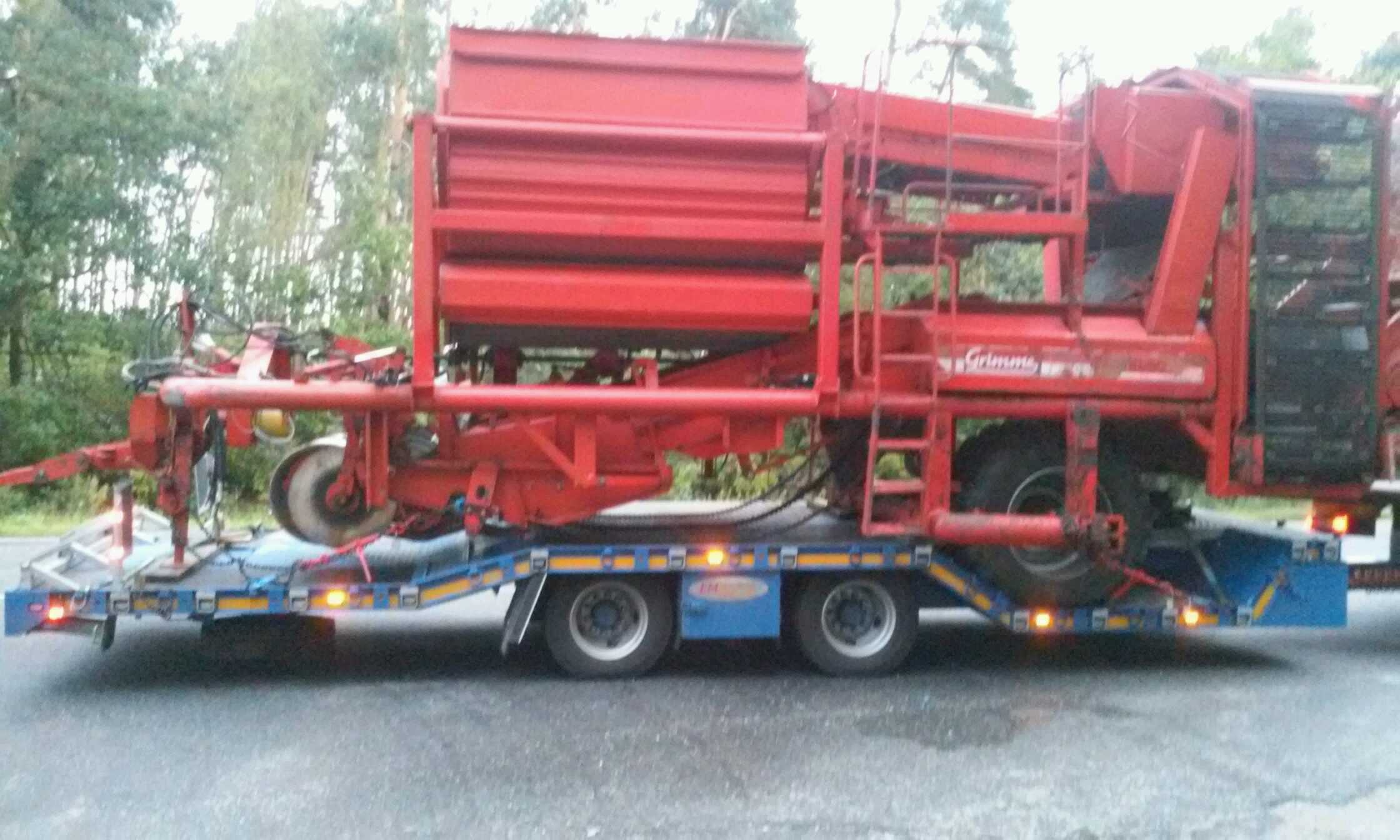 Czerwona maszyna rolnicza Grimme na niskopodwoziowej lawecie z włączonymi światłami pozycyjnymi, transportowana po asfaltowej drodze.