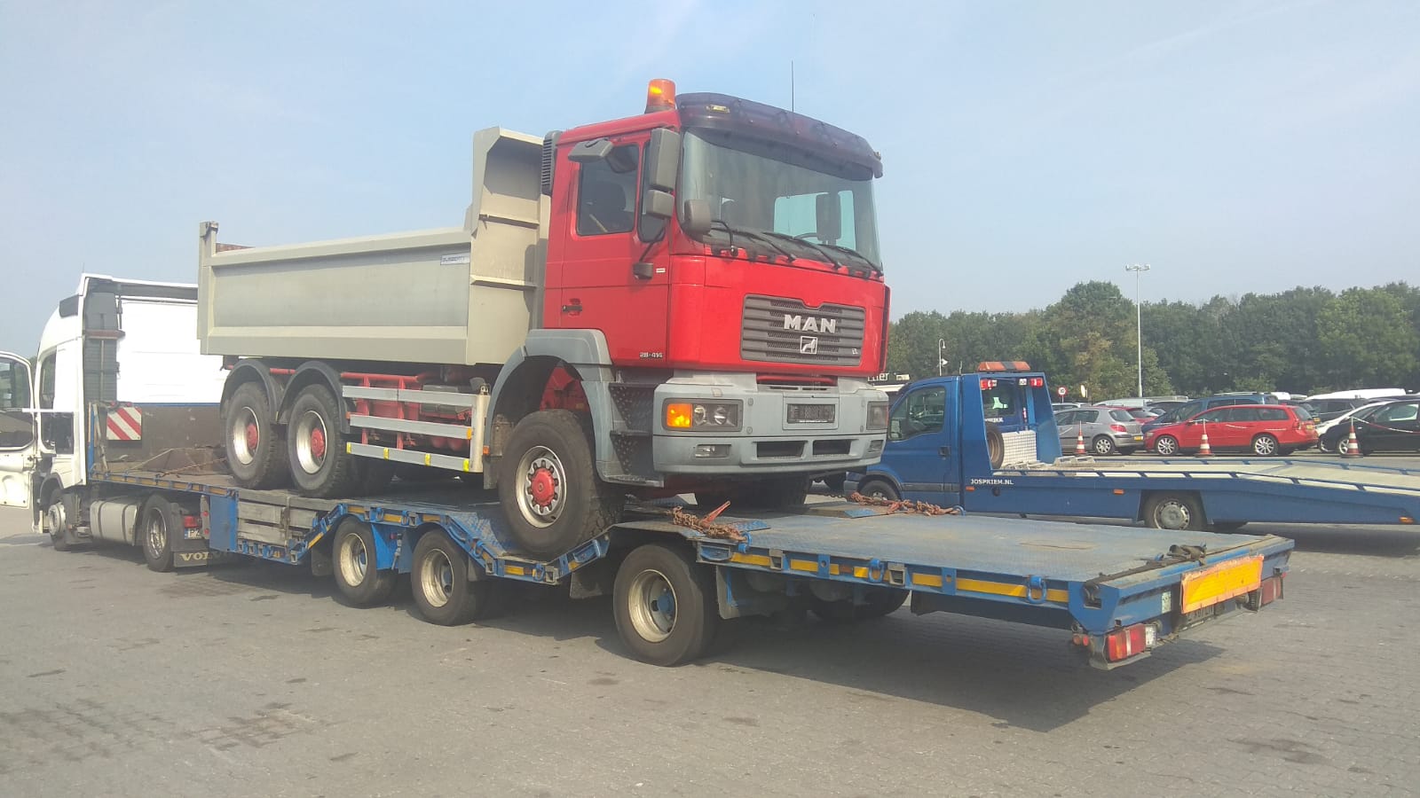 Czerwony samochód ciężarowy MAN z szarą skrzynią ładunkową, zabezpieczony łańcuchami, transportowany na niebieskiej niskopodwoziowej platformie lawety. W tle inny pojazd lawety i parking.