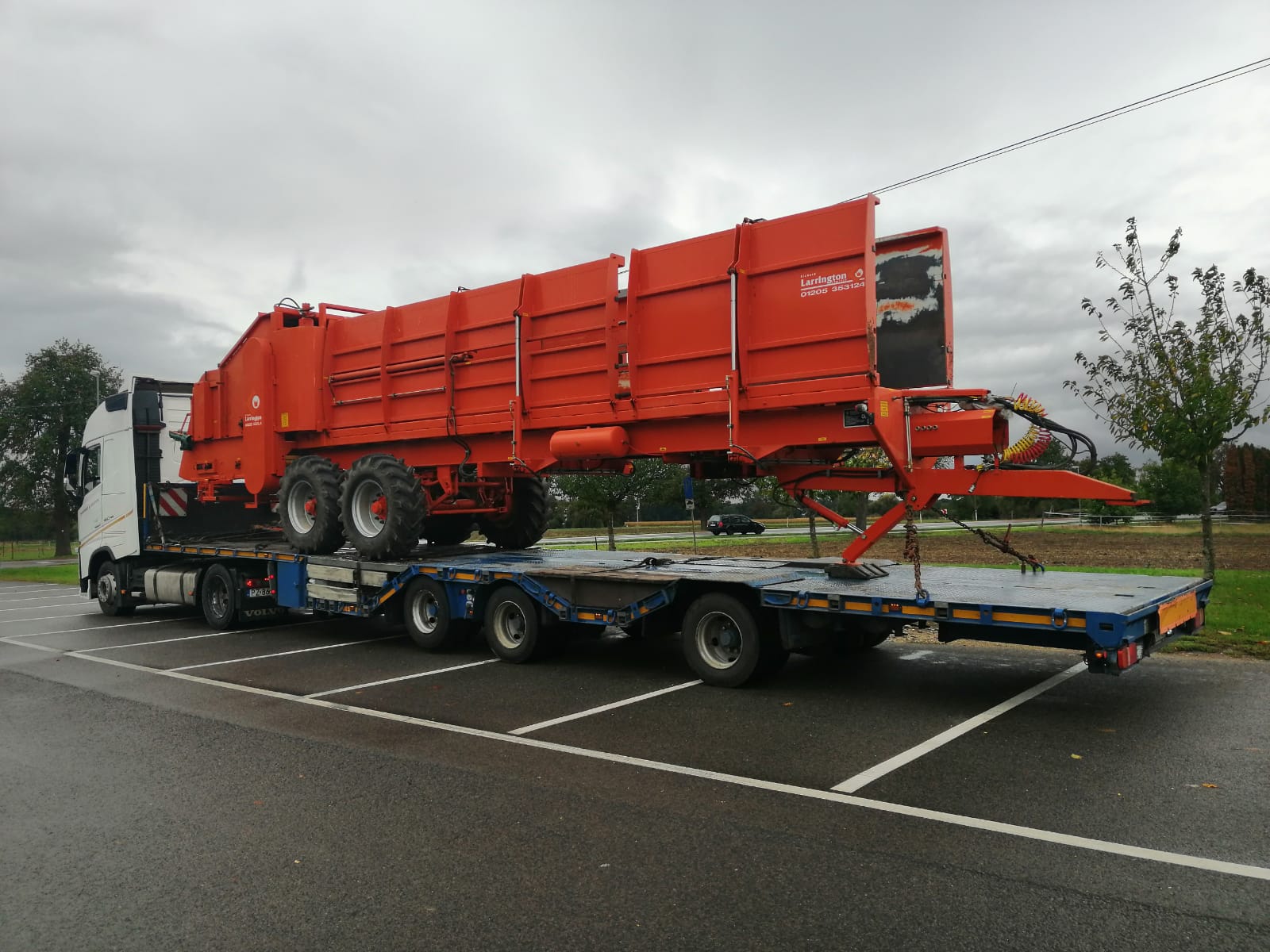 Pomarańczowy rozrzutnik obornika Larrington na niebieskiej lawecie transportowany przez białą ciężarówkę na parkingu w pochmurny dzień.