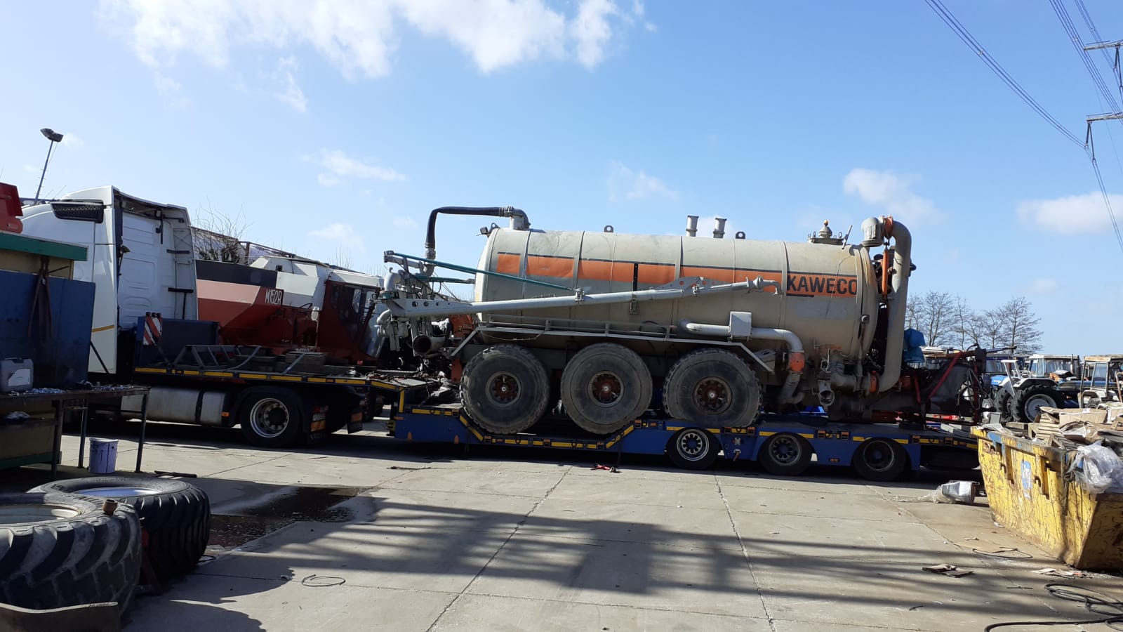 Ciężarówka z naczepą niskopodwoziową przewożąca cysternę KAWECO z wielkimi oponami, widoczne elementy mocowania ładunku, tło: błękitne niebo i słupy wysokiego napięcia.