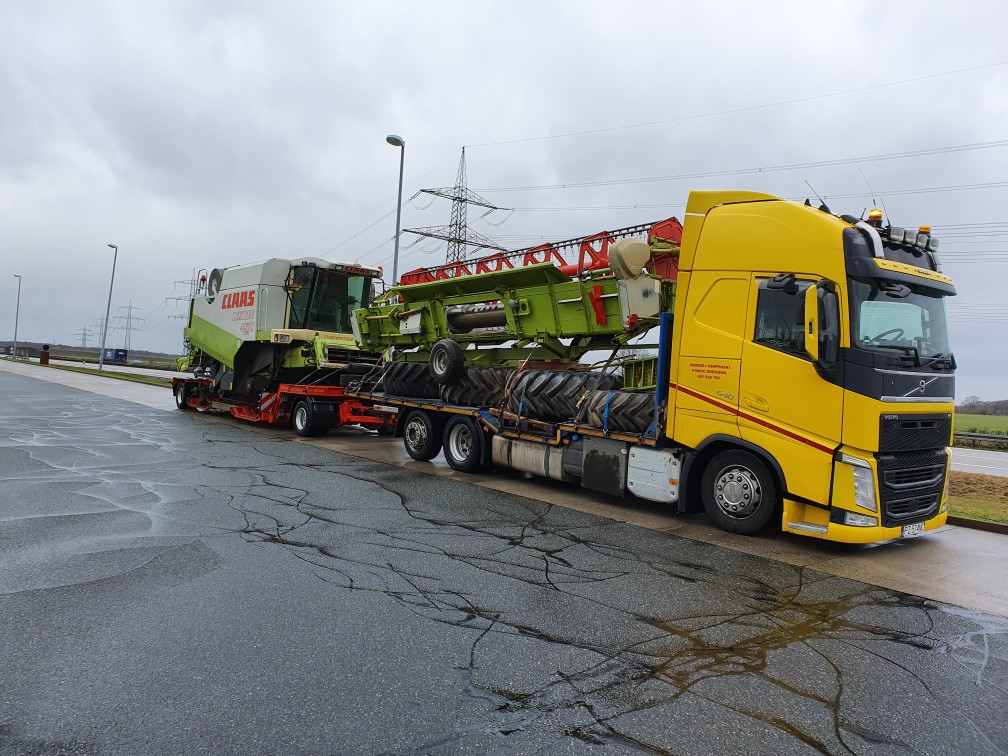 Żółta ciężarówka Volvo przewozi na niskopodwoziowej naczepie kombajn zbożowy Claas i zielony zespół żniwny. Widoczna mokra nawierzchnia parkingu i słupy wysokiego napięcia w tle.