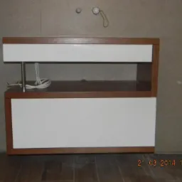 Biała szafka łazienkowa z drewnianym obramowaniem, widoczne przewody i rurka odpływowa, na tle płytek ceramicznych.