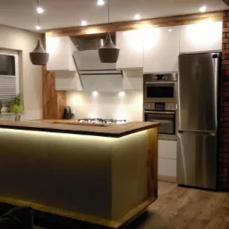 Nowoczesna kuchnia z wyspą oświetloną ledami, białymi szafkami, drewnianym blatem i ceglanym elementem na ścianie.