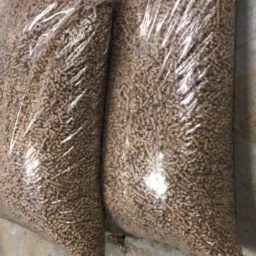 Dwa worki pelletu drzewnego w przezroczystych workach foliowych, leżące na betonowej podłodze obok metalowego wiadra.