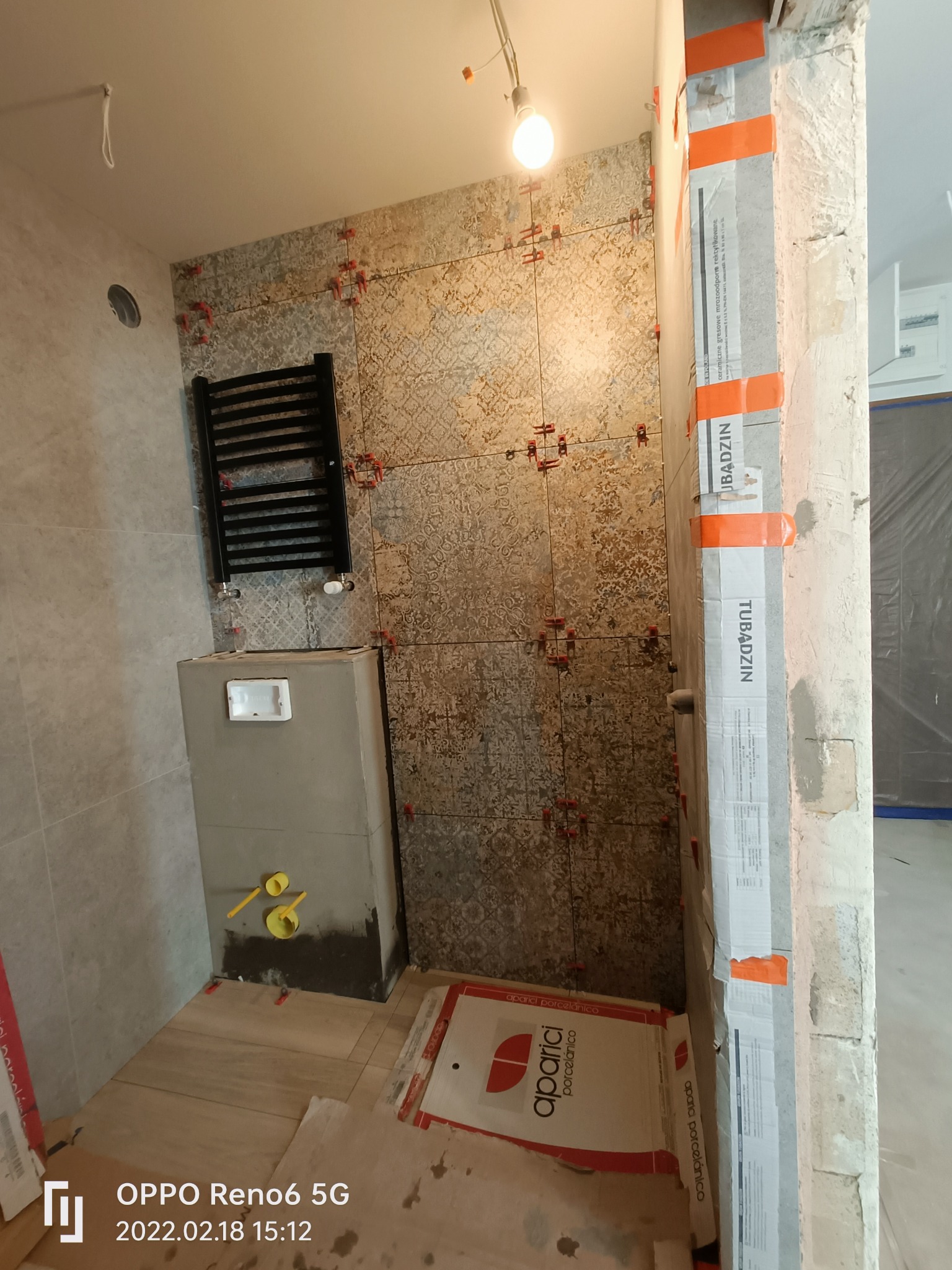 Remont łazienki w trakcie: układanie płytek z wzorem na ścianie, widoczne krzyżyki dystansowe, zamontowany grzejnik drabinkowy, zabudowa stelaża WC, rury z żółtymi zaślepkami, kartony po płytkach...