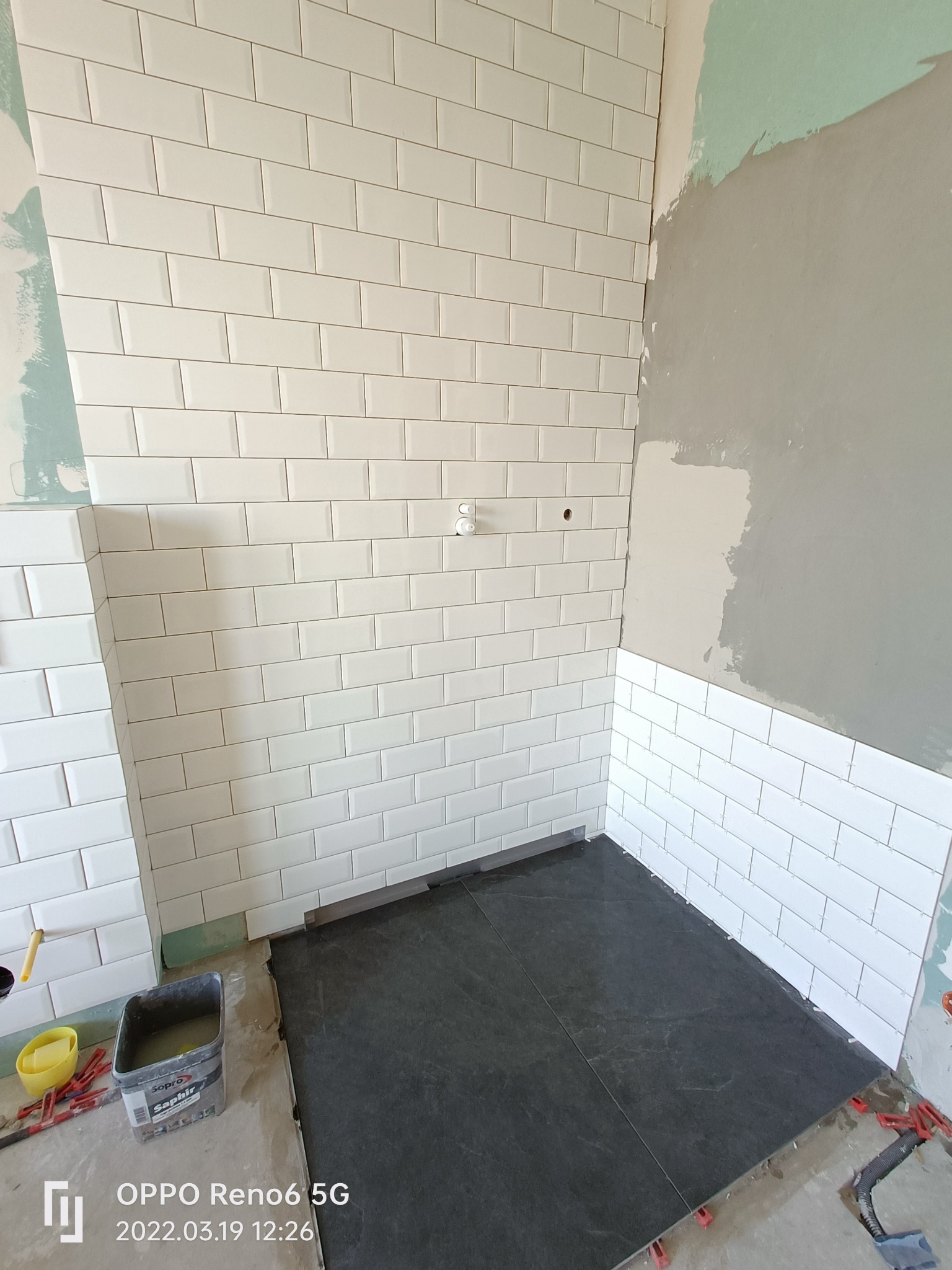 Ściana w trakcie układania białych, prostokątnych płytek typu metro w biurze, widoczne czarne płytki podłogowe i narzędzia glazurnicze.