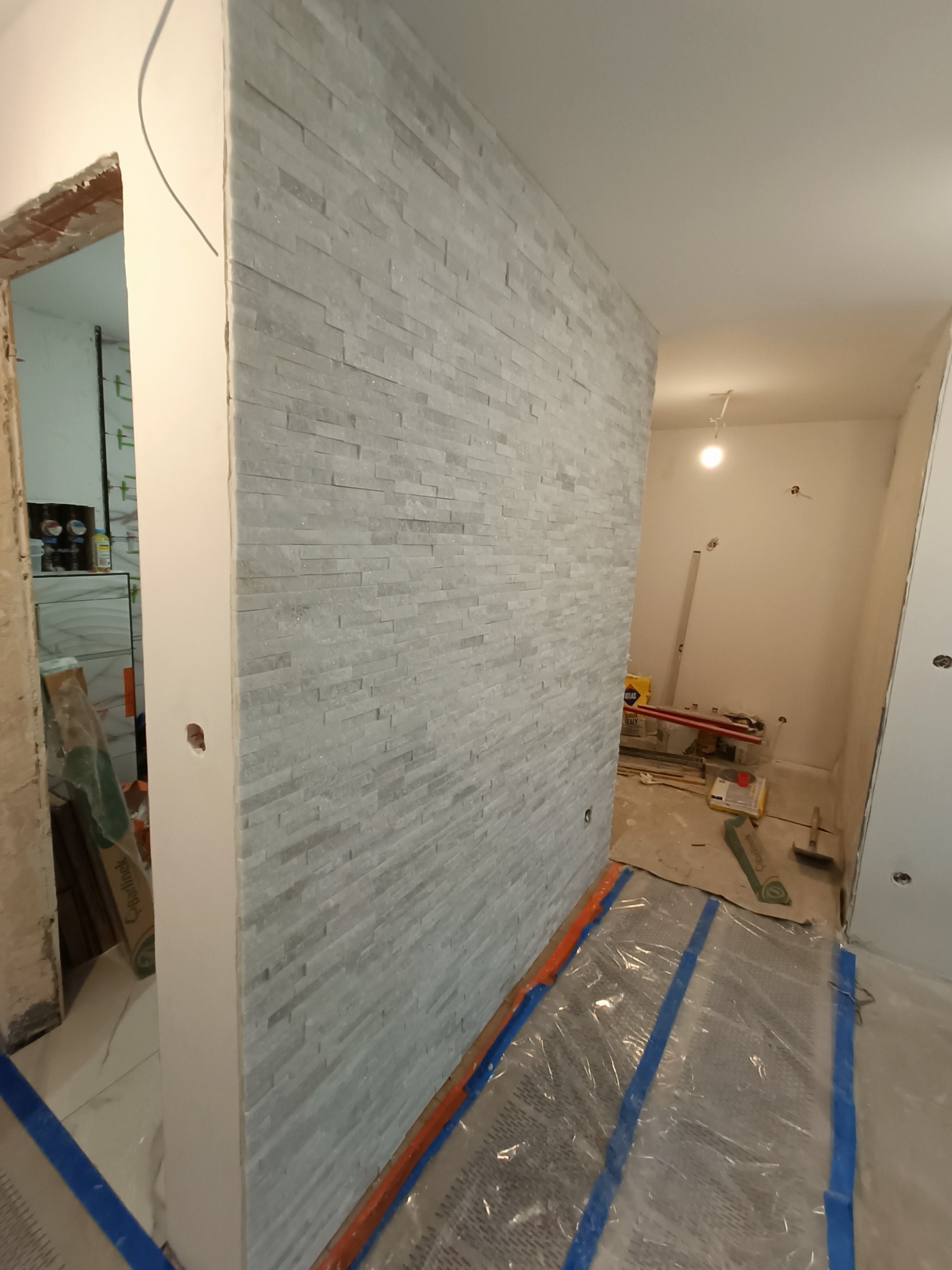 Montaż dekoracyjnej ściany z kamienia naturalnego w biurze, widoczne zabezpieczenia podłogi folią i taśmą, surowe wykończenie ściany po lewej stronie.