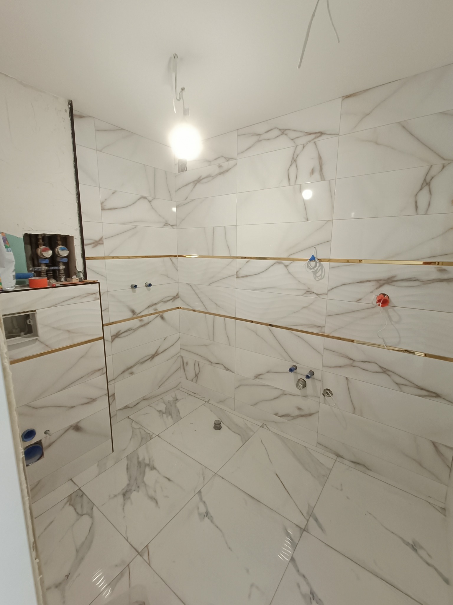 Wnętrze łazienki w trakcie remontu, wyłożone białymi płytkami imitującymi marmur z poziomymi, złotymi akcentami, widoczne przygotowane instalacje wodne i elektryczne, surowy stan wykończenia.