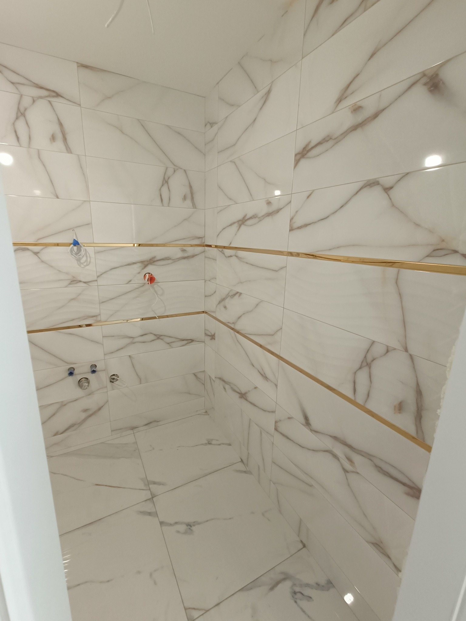 Wykończona łazienka w biurze z białymi płytkami imitującymi marmur z brązowymi żyłami, ozdobnymi złotymi listwami i widocznymi przyłączami hydraulicznymi.