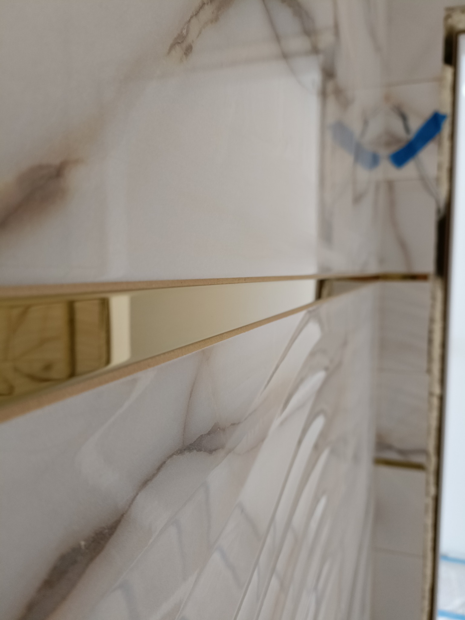 Fragment ściany z ułożonymi białymi płytkami imitującymi marmur, z dekoracyjną złotą listwą, widoczne niebieskie taśmy zabezpieczające.