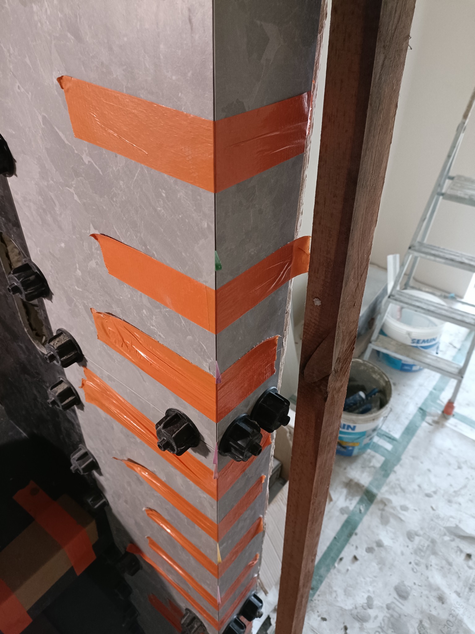 Montaż płytek imitujących kamień na rogu ściany, zabezpieczonych pomarańczową taśmą i czarnymi klipsami poziomującymi, widoczny drewniany element konstrukcyjny i drabina w tle.