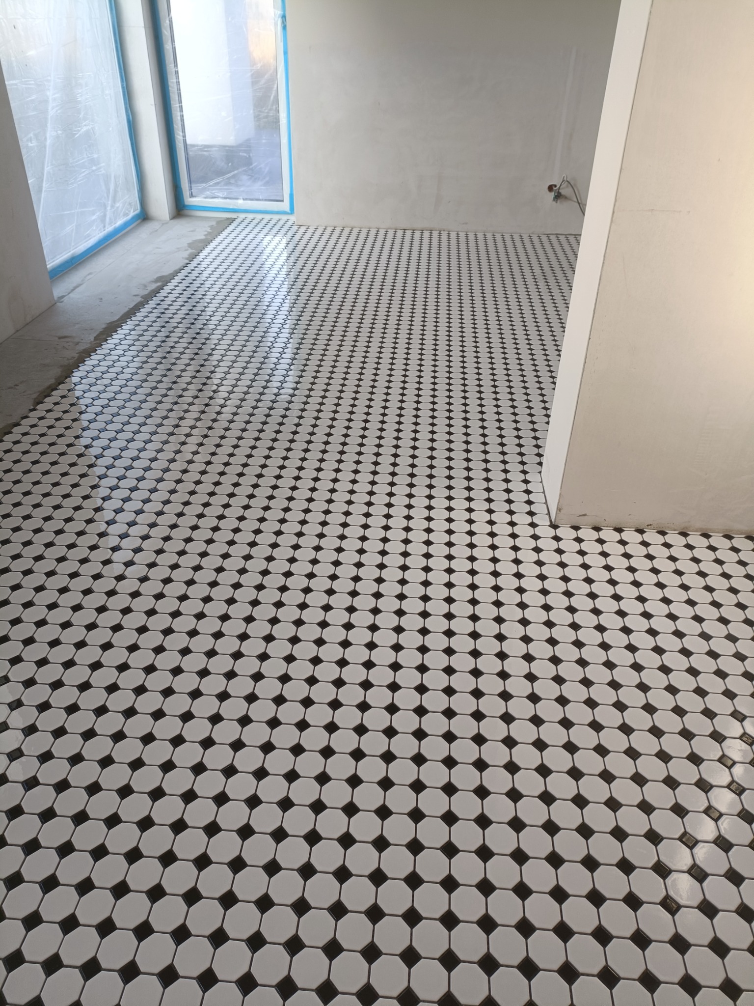 Wykończona podłoga w biurze z białych, ośmiokątnych płytek z czarnymi kwadratowymi wstawkami, widoczne ślady po remoncie na ścianach i folia ochronna na oknie.