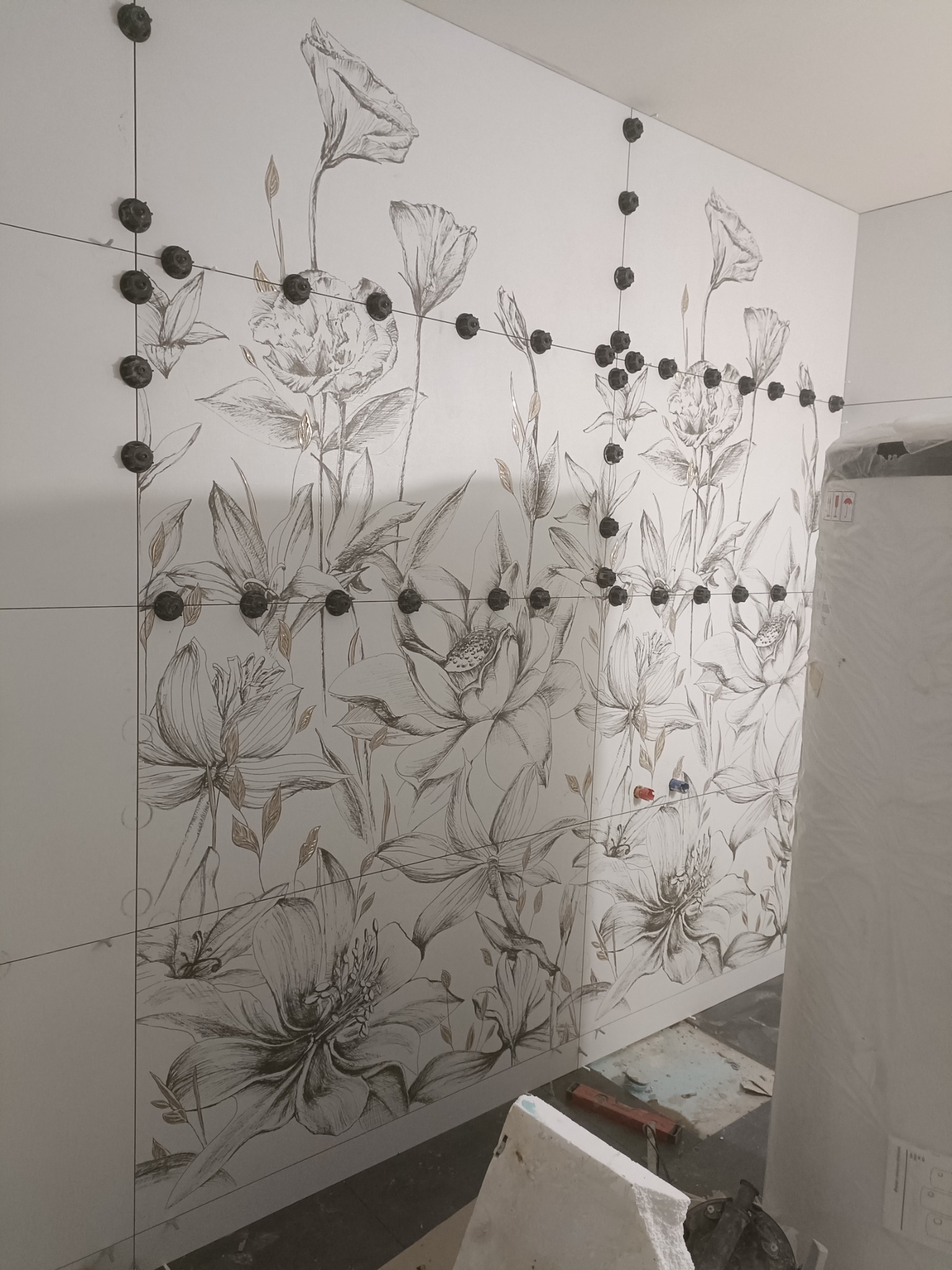 Ściana w trakcie układania płytek z motywem kwiatowym, widoczne czarne klipsy poziomujące i cienkie krzyżyki dystansowe, w tle fragment bojlera.