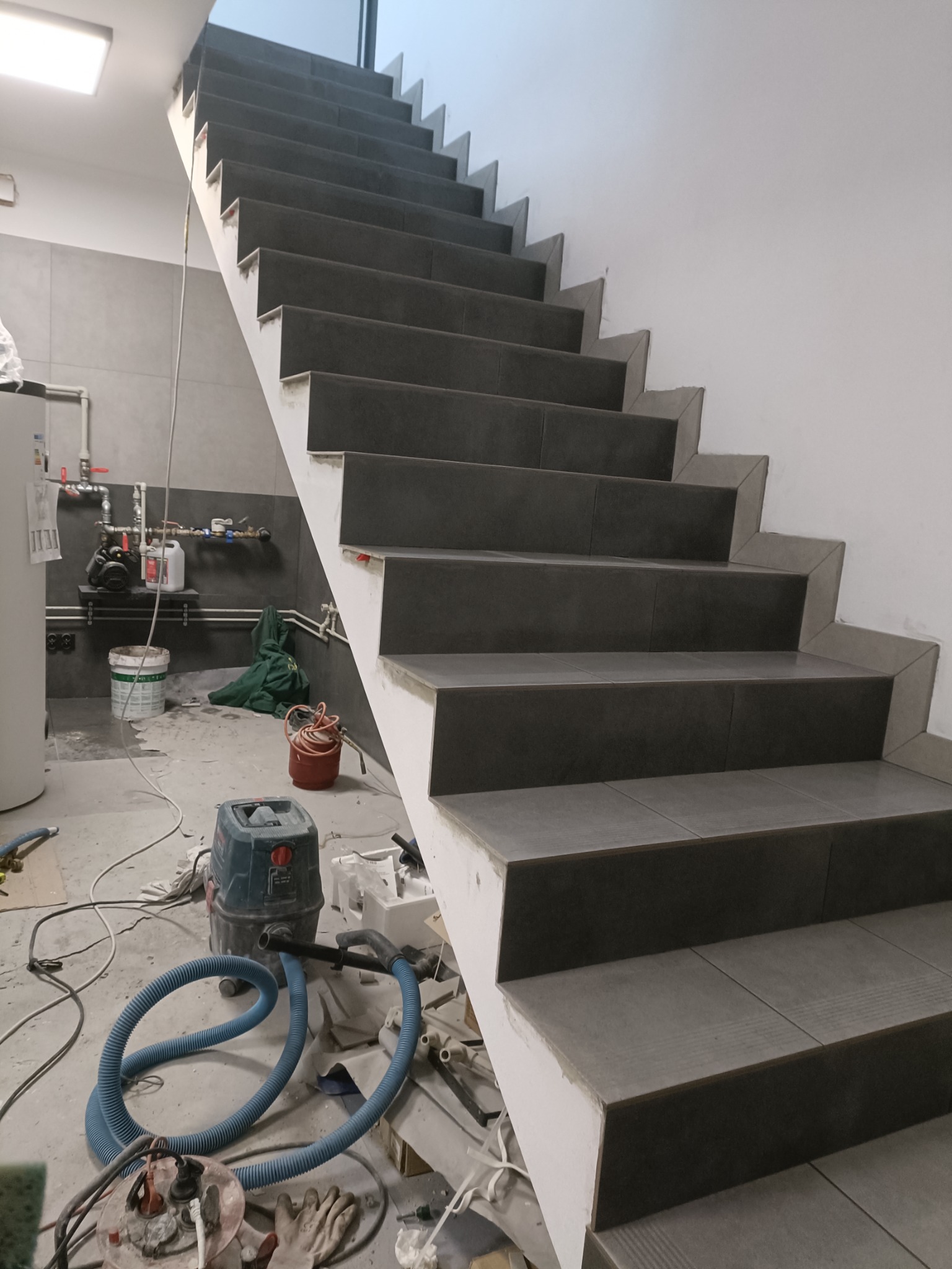 Wykończone schody z szarych płytek w trakcie montażu, widoczny odkurzacz przemysłowy i narzędzia na podłodze, instalacja grzewcza w tle.