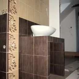 Wykończona łazienka z umywalką nablatową na postumencie z brązowych płytek imitujących drewno, ściany wyłożone beżowo-brązowymi płytkami z motywem roślinnym, widoczne niedociągnięcia wykończeniowe.