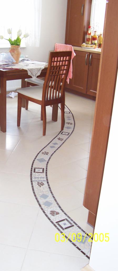 Wykończona podłoga w kuchni z białych płytek z dekoracyjnym, mozaikowym pasem w kształcie łuku, zawierającym motywy kwiatowe i geometryczne; widoczne krzesło i fragment zabudowy kuchennej.