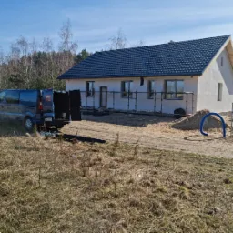 Nowo wybudowany, parterowy dom z ciemną dachówką, częściowo otoczony rusztowaniem, z firmowym vanem z otwartymi drzwiami bagażnika na pierwszym planie.