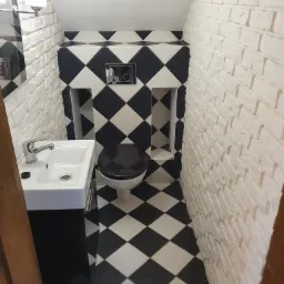 Mała łazienka w stylu retro z białą cegłą na ścianach, czarno-białą szachownicą na podłodze i ścianie za toaletą, oraz umywalką na szafce.