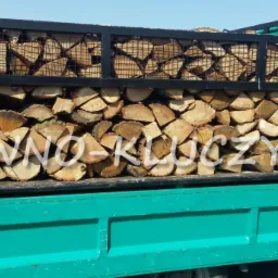 Załadunek pociętego, jasnego drewna w metalowej klatce na platformie turkusowej ciężarówki z napisem 'DREWNO-KLUCZYNSKI'.