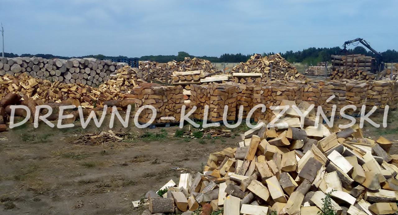 Składowisko pociętego drewna ułożonego w stosy, z napisem 'DREWNO-KLUCZYŃSKI' na pierwszym planie, w tle widoczny dźwig do załadunku.