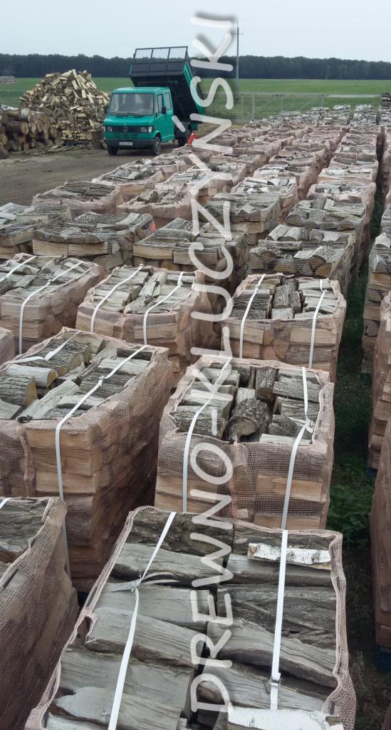 Ułożone w rzędach, zapakowane w siatkę i obwiązane taśmą paczki drewna kominkowego na placu składowym, w tle zielona ciężarówka i sterta drewna luzem.