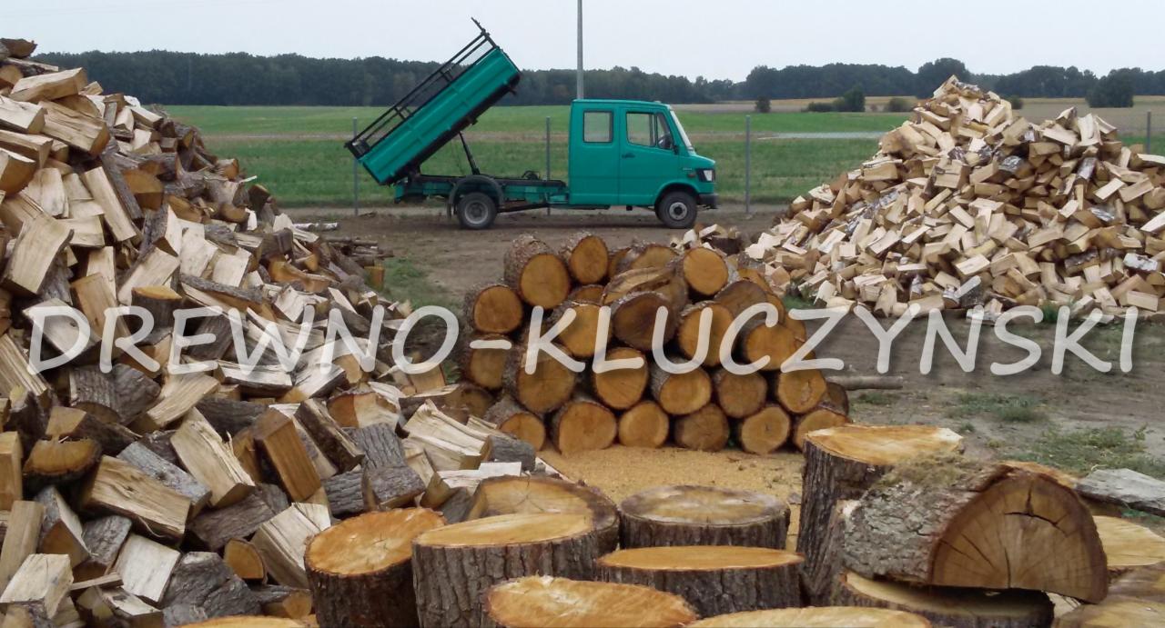 Stosy pociętego drewna ułożone obok zielonej ciężarówki z podniesioną skrzynią ładunkową na tle pola i lasu.