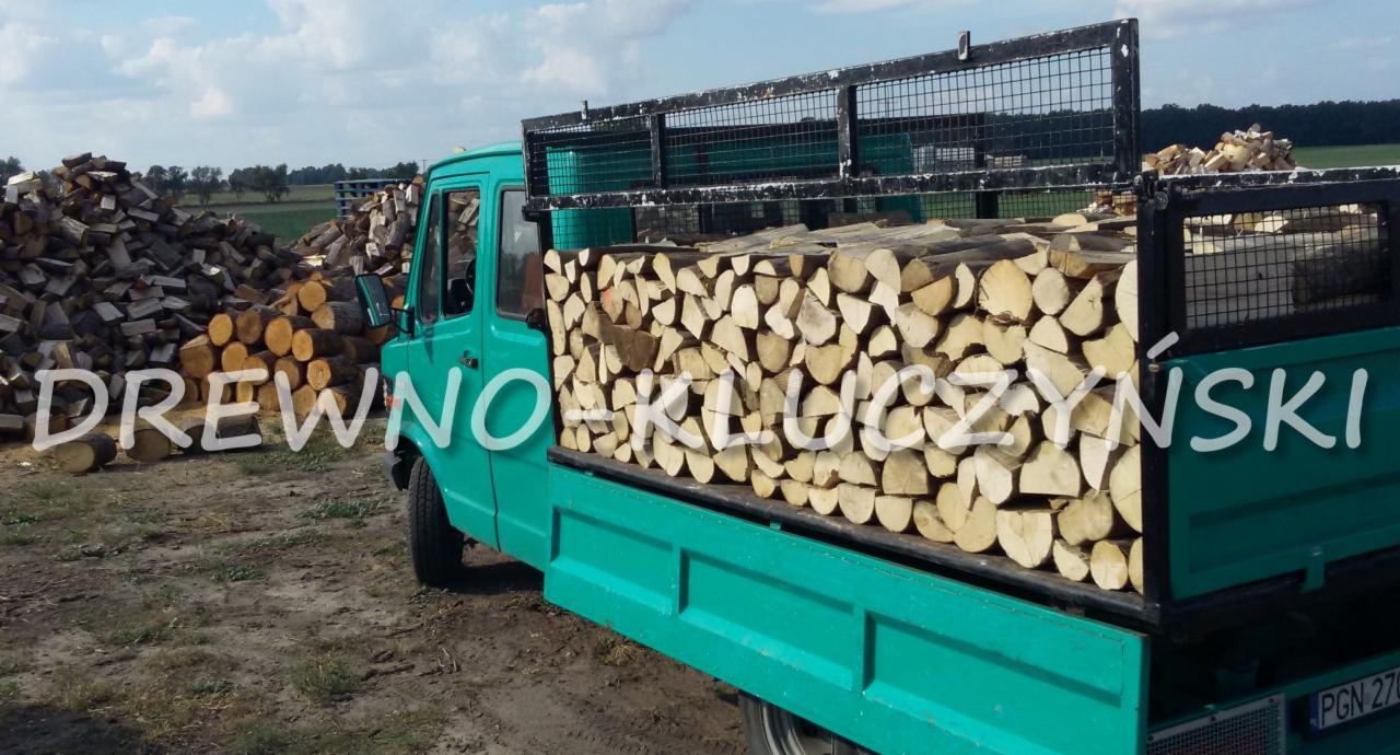 Turkusowa ciężarówka z metalową klatką wypełniona pociętymi kawałkami drewna, z tyłu widoczny stos drewna na polu.