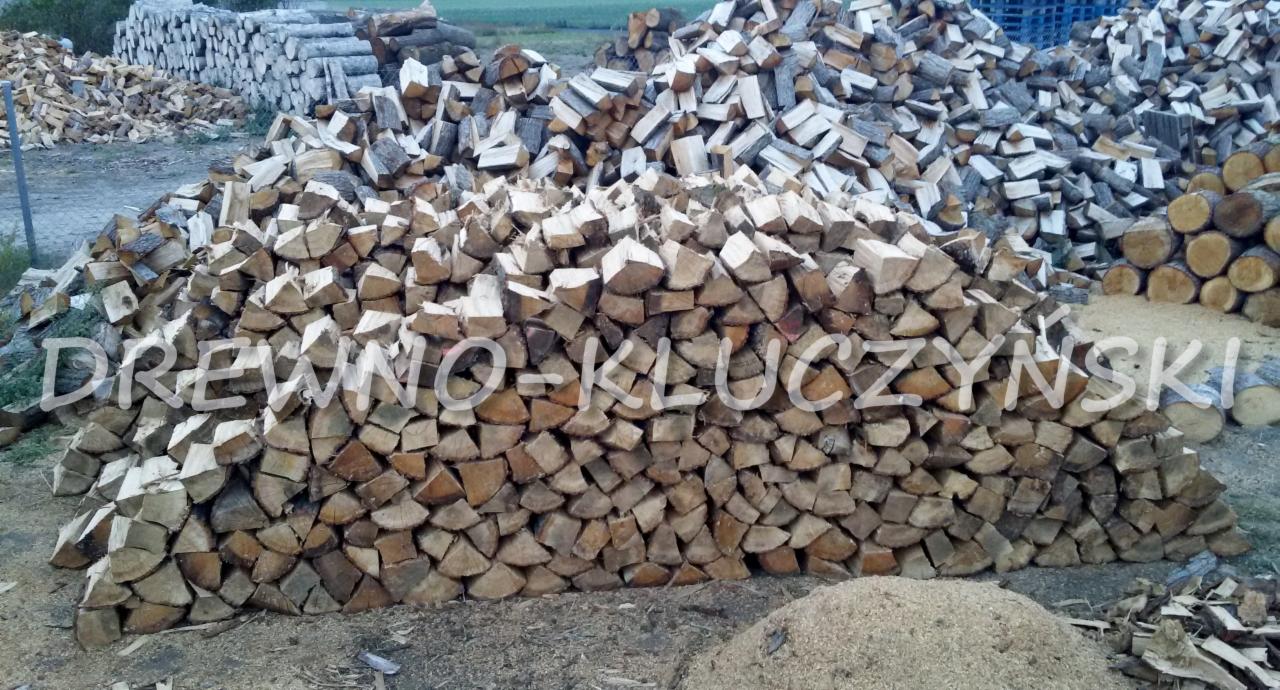 Stos pociętego drewna ułożony w pryzmę, z widocznymi okrąglakami w tle i stertą trocin u podstawy.