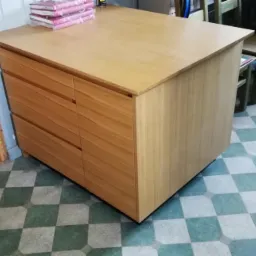 Jasne, drewniane biurko z szufladami i blatem roboczym, ustawione na podłodze w szachownicę. Na blacie leżą książki lub notesy w różowych okładkach.