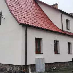 Odnowiona elewacja budynku z czerwonym dachem, widoczna antena satelitarna i TV, kamienne fundamenty oraz drewniane zadaszenie nad wejściem.