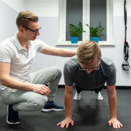 Mężczyzna w okularach wykonuje pompki na czarnej macie pod nadzorem mężczyzny w okularach, który kuca obok i trzyma rękę na jego plecach; widoczne rośliny w doniczkach i sprzęt do ćwiczeń w tle.