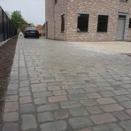 Nowo ułożona, szara kostka brukowa prowadząca do ceglanego domu z czarnymi oknami, widoczny czarny samochód zaparkowany w oddali.