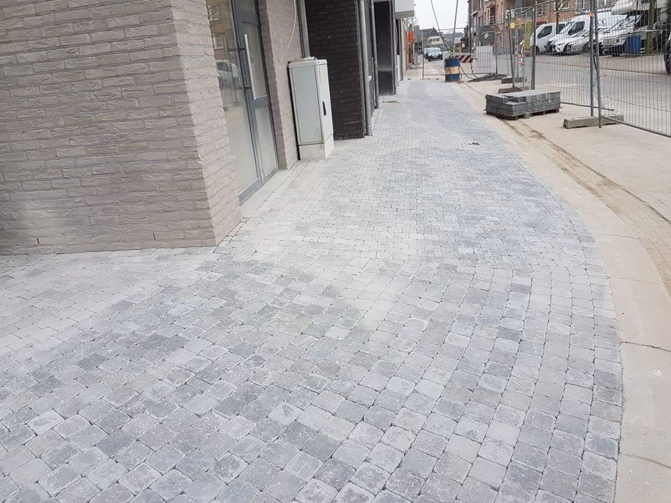 Nowo ułożona szara kostka brukowa na chodniku przed budynkiem z cegły, widoczne palety z kostką i barierki zabezpieczające.