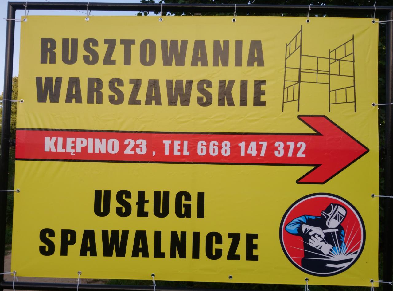Żółty baner reklamowy z napisem 'Rusztowania Warszawskie', adresem 'Kłępino 23' i numerem telefonu, strzałką kierunkową oraz grafiką spawacza i napisem 'Usługi Spawalnicze'.