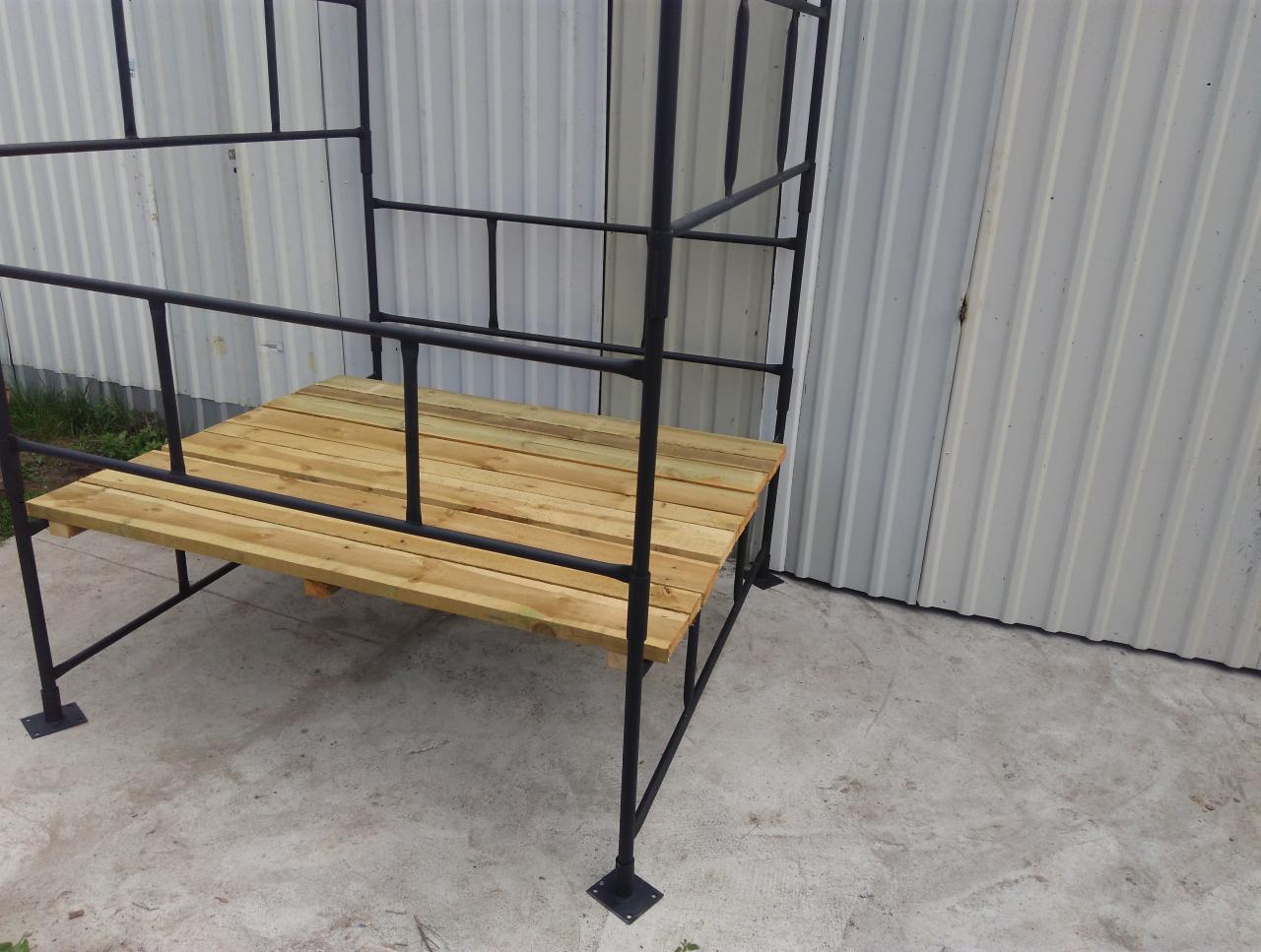 Konstrukcja z czarnych rur i surowego drewna, prawdopodobnie w trakcie budowy, na tle blaszanej ściany.