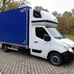 Biały samochód dostawczy Renault Master z niebieską plandeką na parkingu z betonowej kostki, jesienne drzewa w tle.