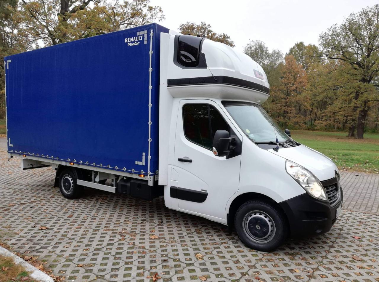 Biały samochód dostawczy Renault Master z niebieską plandeką na parkingu z betonowej kostki, jesienne drzewa w tle.