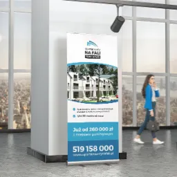 Roll-up reklamowy z wizualizacją osiedla apartamentów 'Apartamenty Na Fali' w Pobierowie, z widoczną przechodzącą kobietą rozmawiającą przez telefon na tle okna z panoramą miasta.