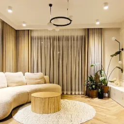 Jasny salon z beżową sofą, okrągłym stolikiem kawowym z lamelami, wzorzystym dywanem i zasłonami w odcieniach beżu i bieli, oświetlony lampą wiszącą z kulistymi kloszami.