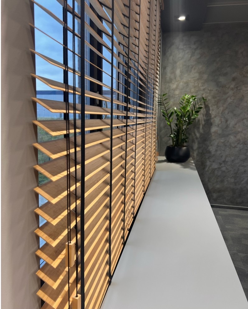 Zamknięte drewniane żaluzje w jasnym odcieniu, czarne sznurki, widok z boku, roślina doniczkowa na parapecie.