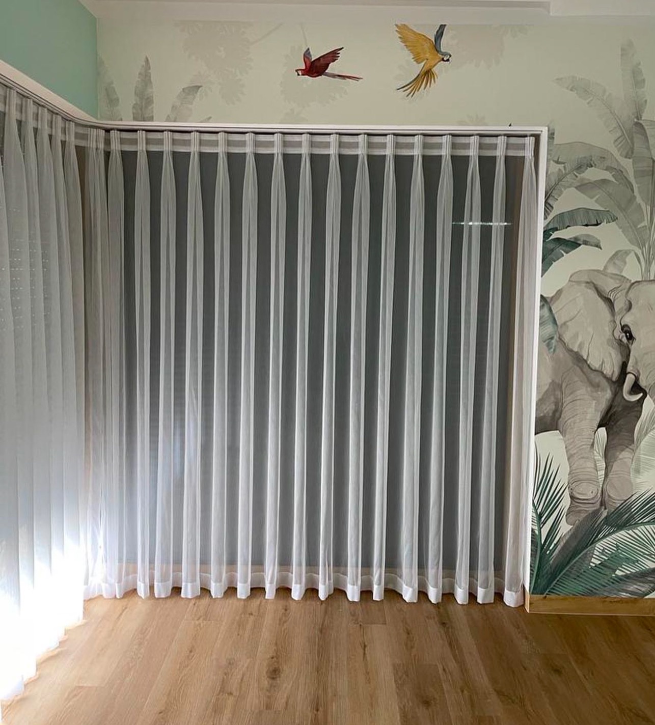 Delikatne, białe, półprzezroczyste zasłony panelowe zawieszone na karniszu szynowym, w tle ściana z motywem dżungli i słoni oraz drewniana podłoga.