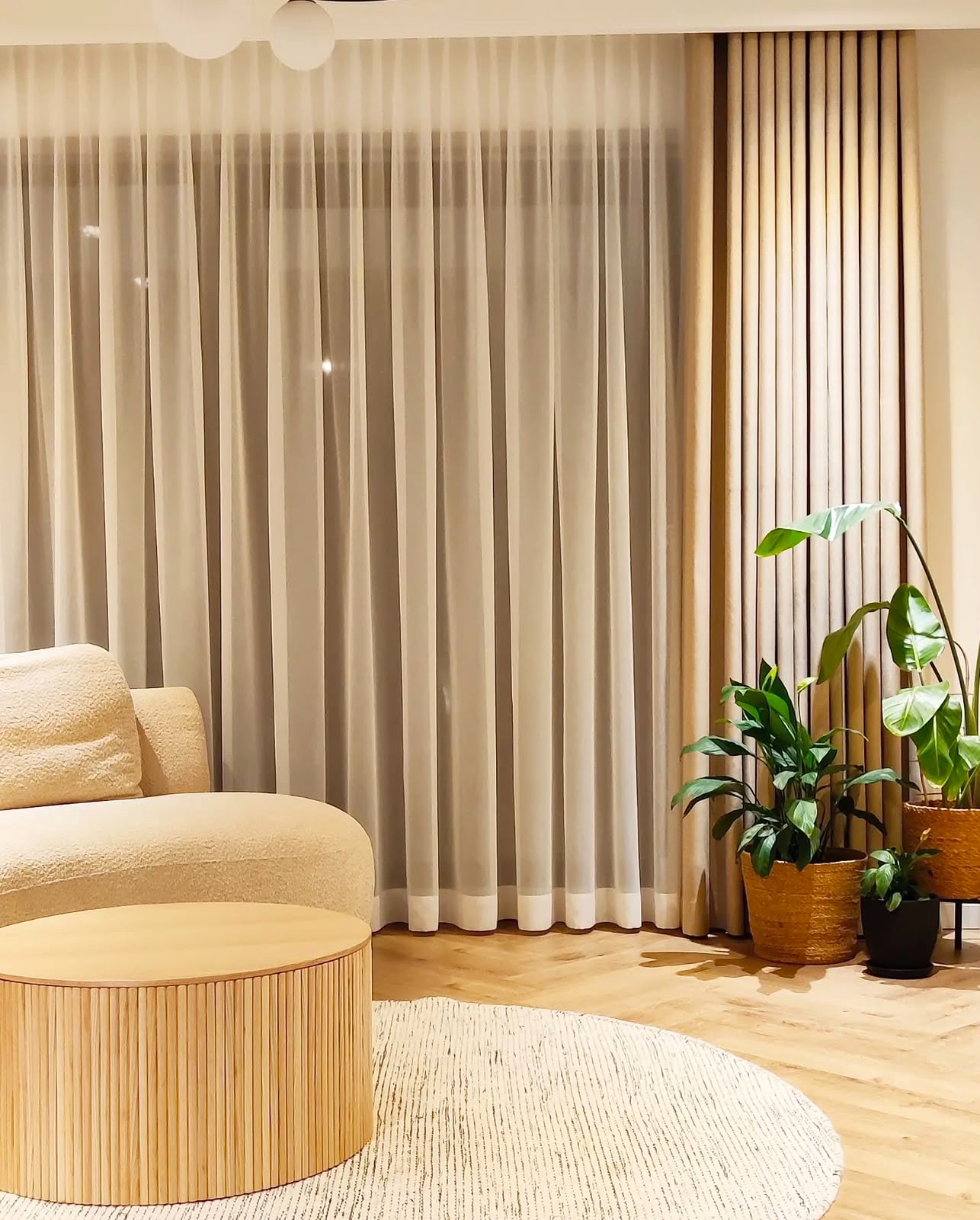 Jasne, przesuwne zasłony z delikatnej tkaniny i grubsze, beżowe zasłony w tle, uzupełnione roślinami doniczkowymi w wiklinowych osłonkach, tworzące przytulne wnętrze salonu z okrągłym stolikiem...