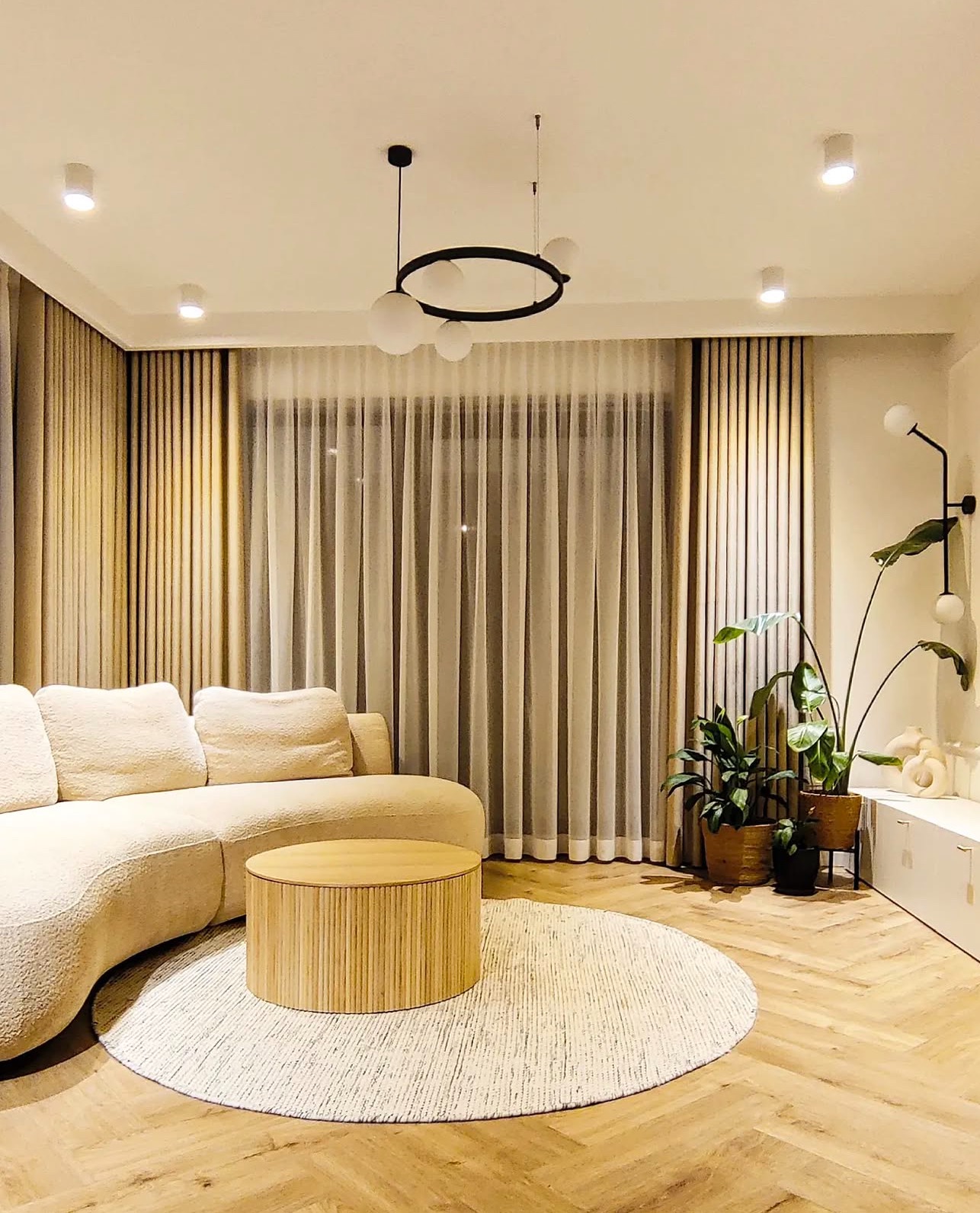 Jasny salon z beżową sofą, okrągłym stolikiem kawowym z lamelami, wzorzystym dywanem i zasłonami w odcieniach beżu i bieli, oświetlony lampą wiszącą z kulistymi kloszami.