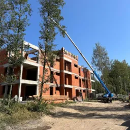 Wznoszenie trzypiętrowego budynku z czerwonej cegły ceramicznej, widoczny dźwig teleskopowy i ułożone deski, teren budowy w słoneczny dzień.