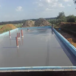 Świeżo wylana, gładka wylewka betonowa w obrysie fundamentów z izolacją, wystające rury instalacyjne, w tle hałdy ziemi i krajobraz.